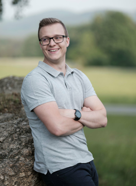 Franz Honeder steht an einen großen Granitstein gelehnt inmitten der schönen Mühlviertler Landschaft und blickt lächelnd in die Ferne.