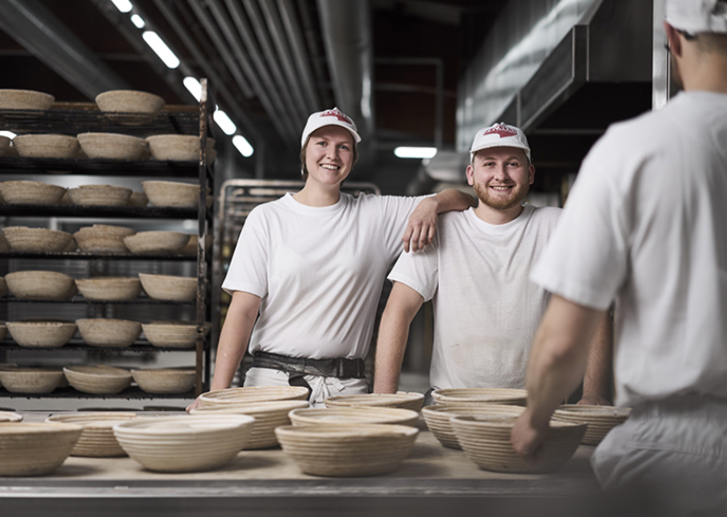 Drei Bäcker stehen vor der Arbeitstafel in der Backstube. Auf der Tafel liegen Brotsimperl. Die Bäckerin legt ihrem Kollegen locker ihren Arm auf seine Schulter. Beide lächeln in die Kamera.