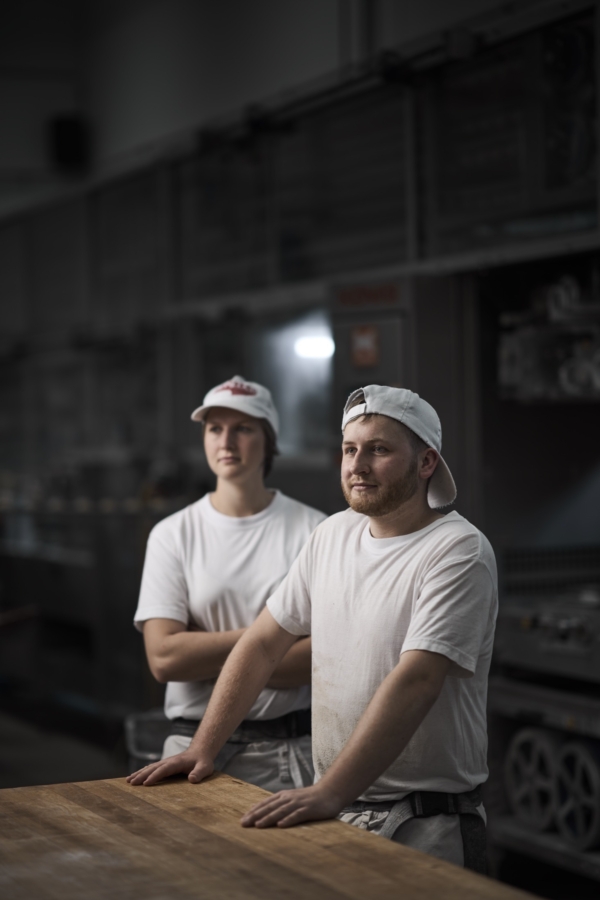 Eine Bäckerin und ein Bäcker stehen in der Honeder Backstube am Wirktisch und blicken in die Backstube