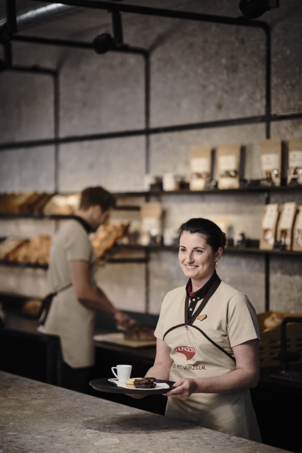 Die Honeder Verkäuferin trägt eine beige Honeder Schürze, hält ein Tablett mit einem Kaffee darauf in der Hand und blickt freundlich in die Ferne