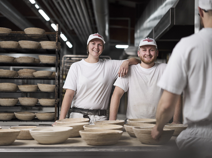 Drei Bäcker stehen vor der Arbeitstafel in der Backstube. Auf der Tafel liegen Brotsimperl. Die Bäckerin legt ihrem Kollegen locker ihren Arm auf seine Schulter. Beide lächeln in die Kamera.